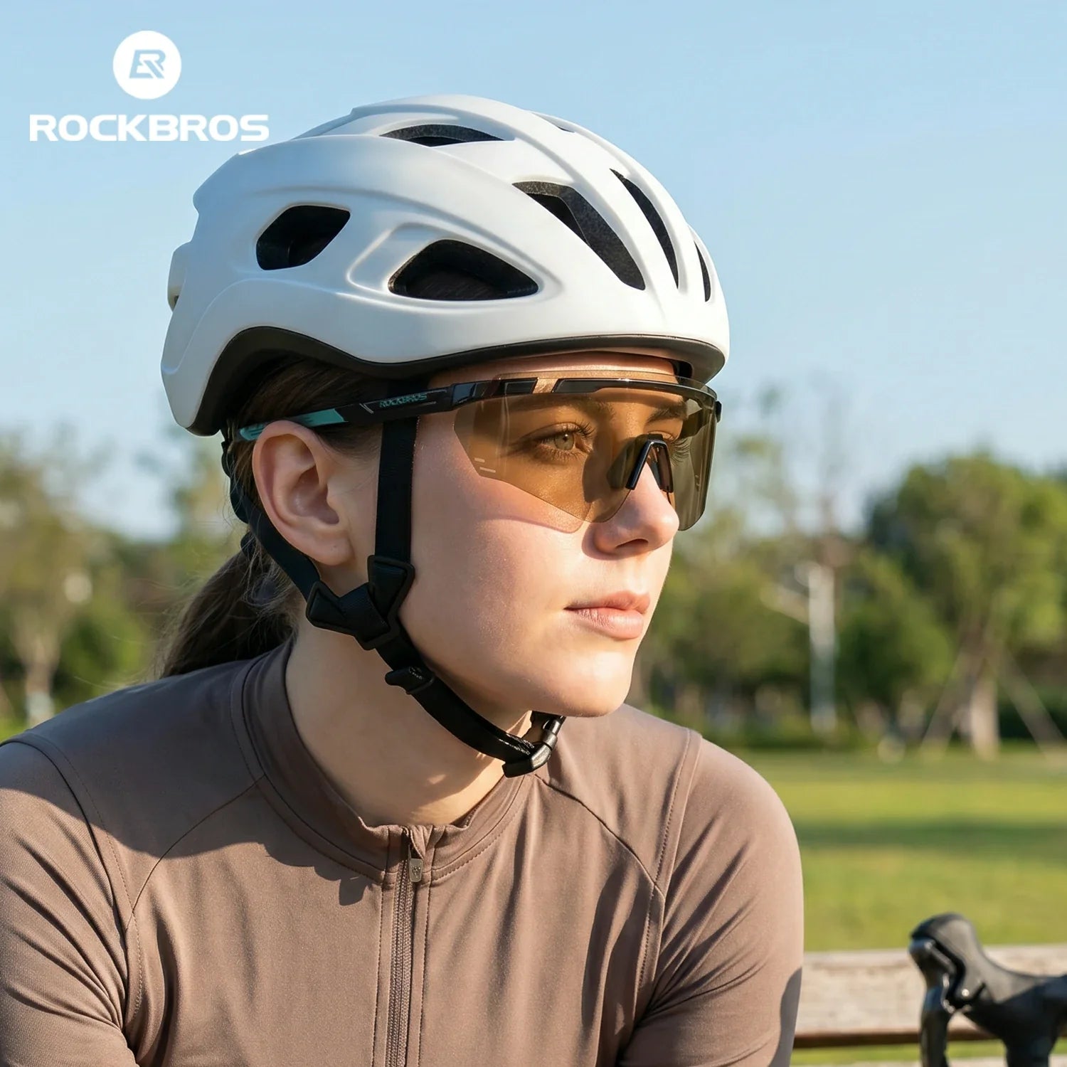 A woman wearing a white Rockbros cycling helmet and tinted sunglasses, dressed in a brown cycling jersey, set against a sunny park background with the Rockbros logo in the top-left corner. #Color_Tea Brown