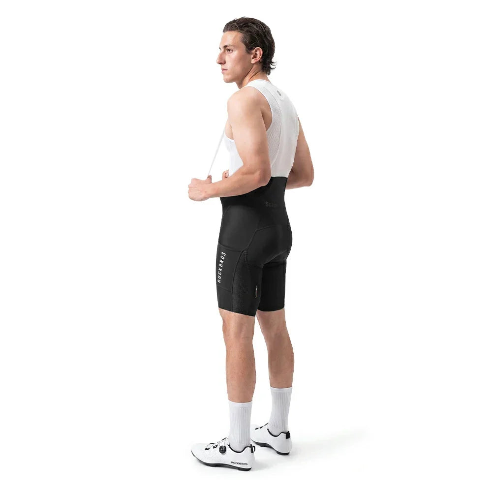 A man in a white cycling jersey and black cycling shorts stands in profile against a clean white background, wearing white cycling shoes and white socks.