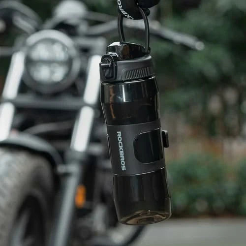 Black water bottle with 'Rockbros' branding hanging from a bike handlebar against a blurred natural background.