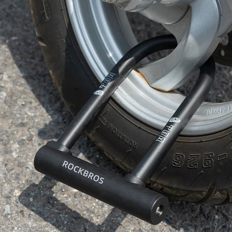 A close-up of a black U-lock bicycle wheel lock branded ROCKBROS, secured around a silver metal wheel rim on a paved ground.