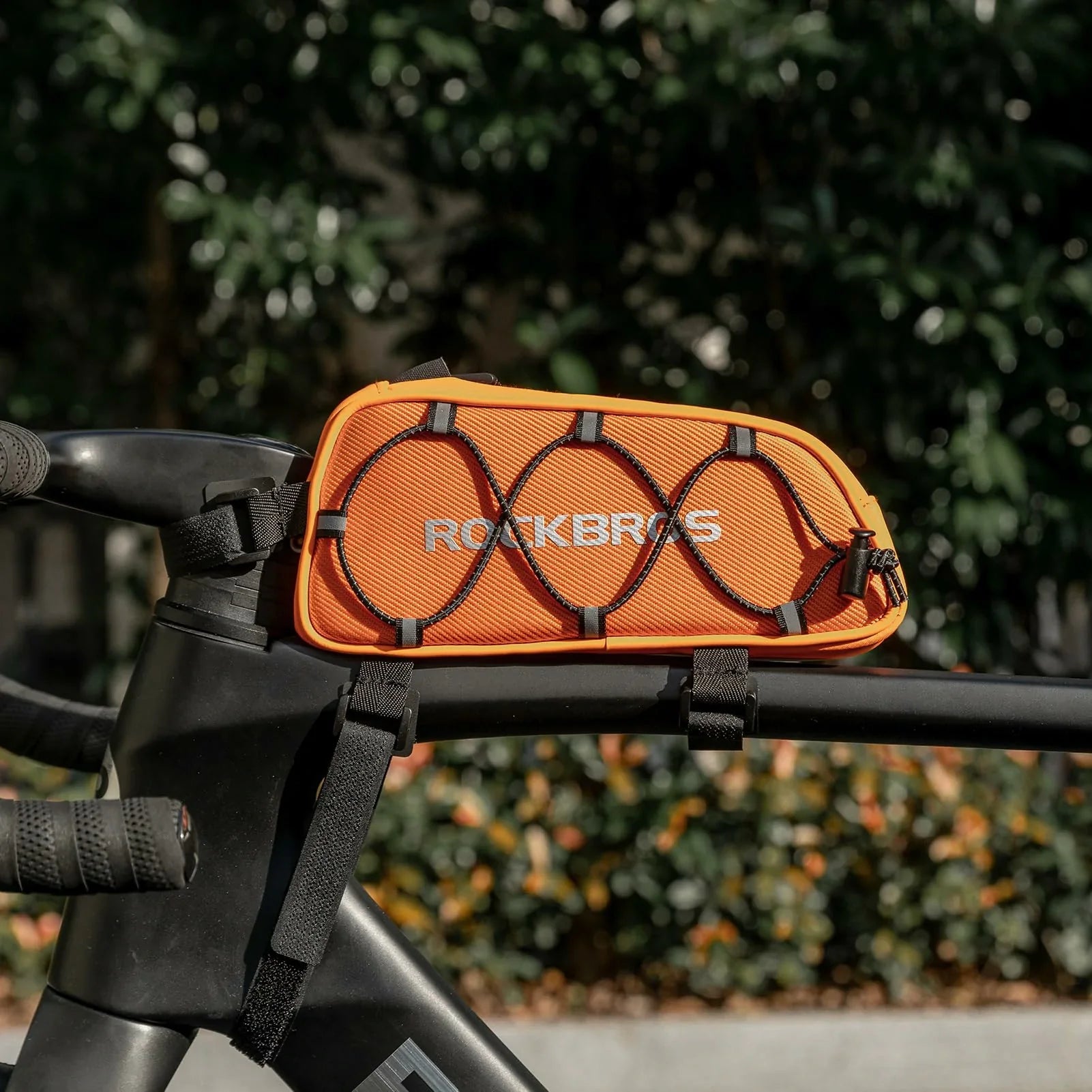 Orange bike bag with 'RockBros' branding attached to a bicycle handlebar, blurred green foliage in the background. #color_orange
