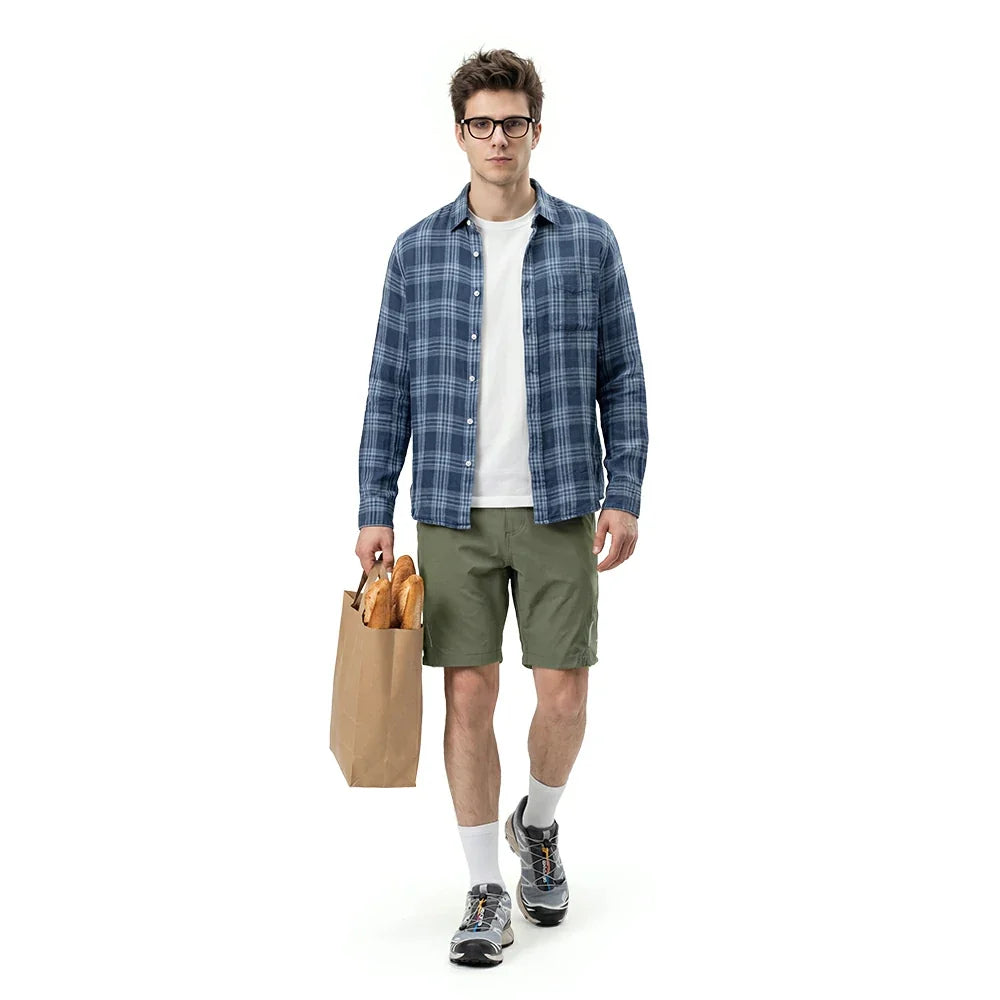 Man wearing a blue plaid shirt, white t-shirt, green shorts, and glasses, holding a paper bag with bread on a white background.