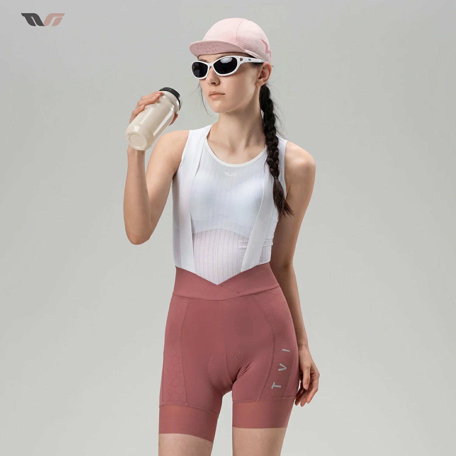 A woman wearing a white ribbed athletic tank top and dusty rose high-waisted cycling shorts, holding a water bottle. She is wearing a pink cap and white sunglasses against a solid light gray studio background.