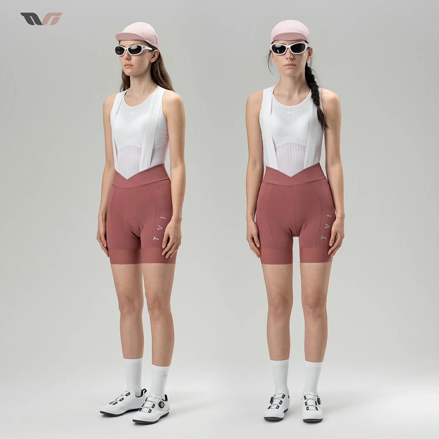 Two female models standing side by side in a studio, wearing matching athletic cycling outfits consisting of white bib shorts and dusty rose fitted shorts, white socks, and cycling shoes. Both wear light pink caps and dark sunglasses against a light gray background.